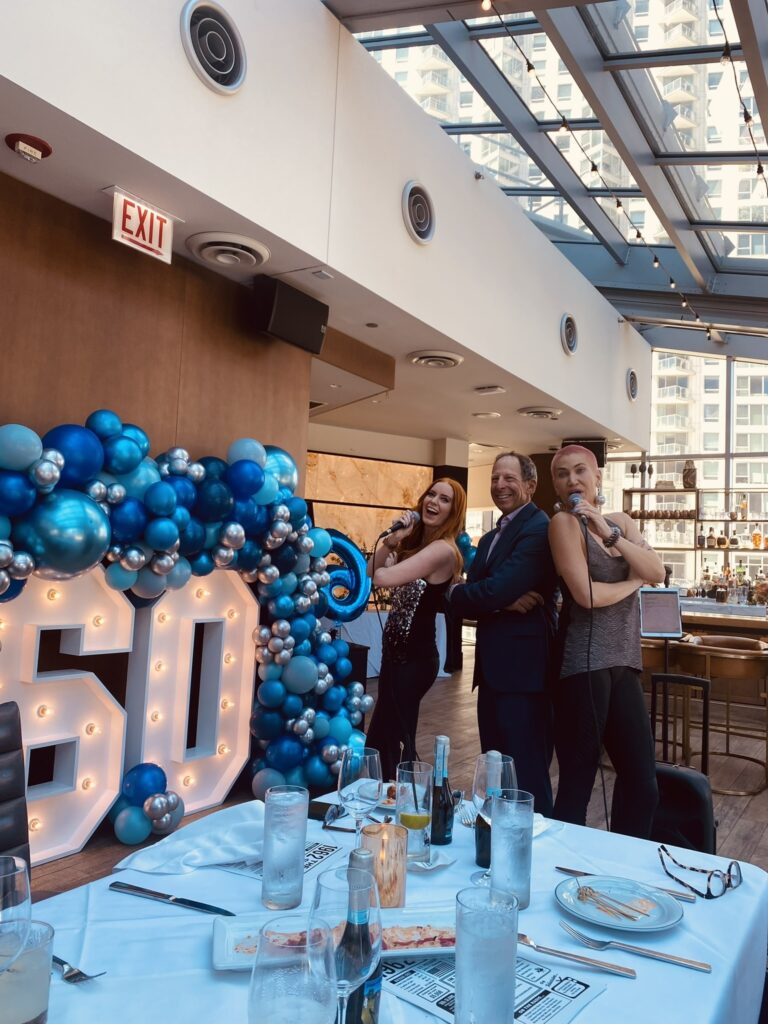 a group of people standing around a table with balloons and a cake