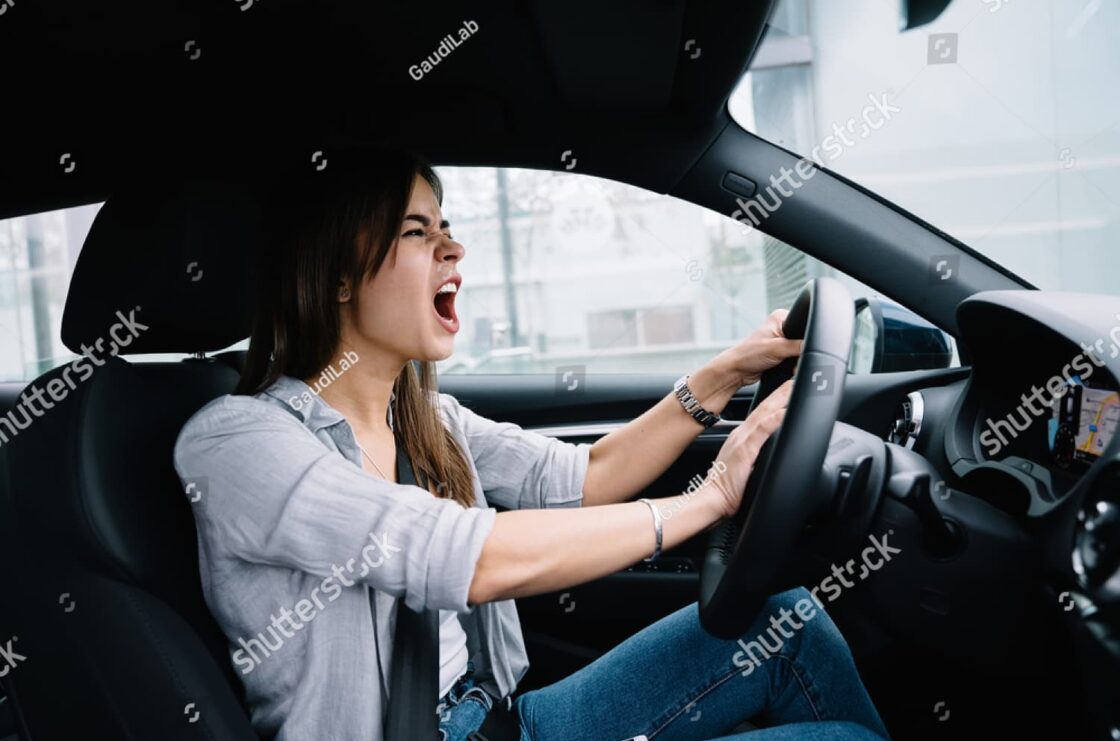 Side view of angry young woman yelling while driving, illustrating verbal aggression as a sign of escalating road rage.